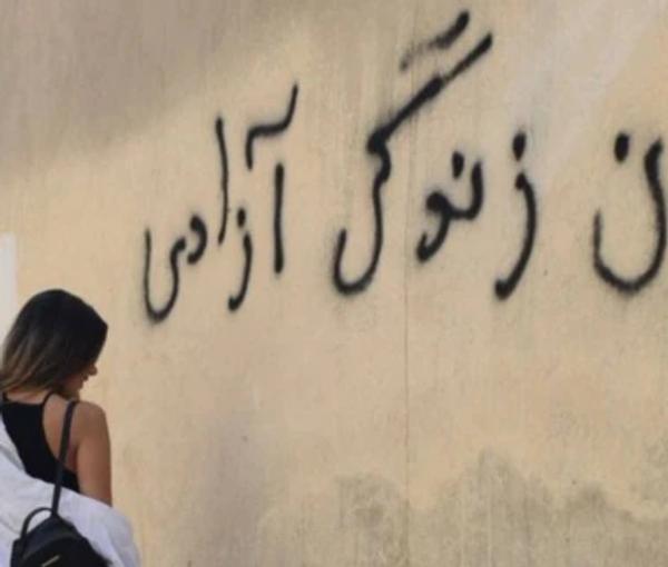 An Iranian woman standing in front of a wall on which the main slogan of the ongoing protests – Women, Life, Liberty – is seen