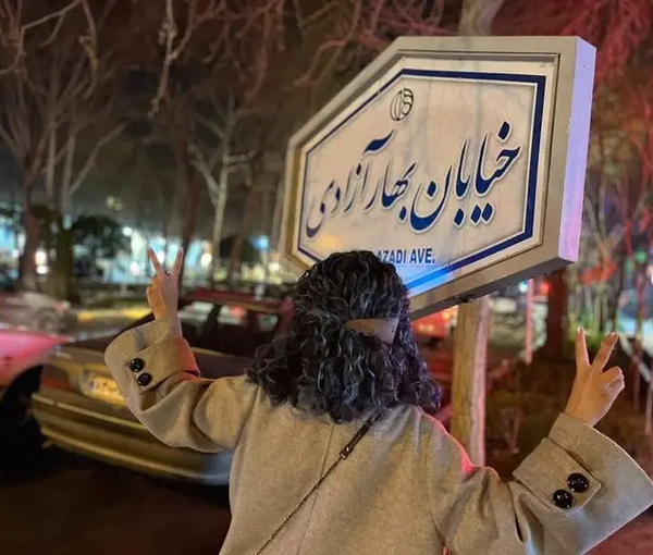 An Iranian woman unveiling in public in front of a street sign that reads “the spring of freedom street” in Tehran (March 13, 2023) 